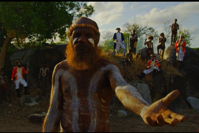 An Aboriginal man in ceremonial paint gestures towards the camera