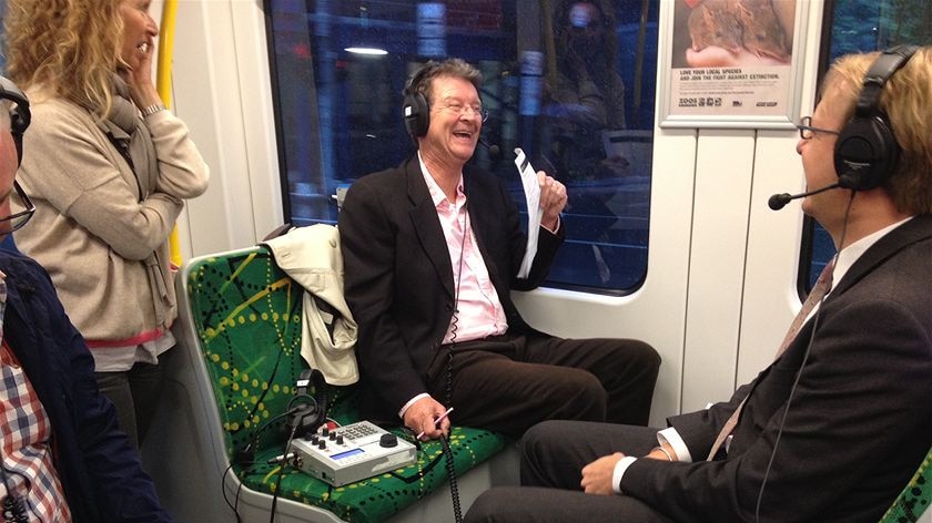 A man laughs as he sits on a tram, wearing a radio headset. Person wearing headset sits opposite, people standing nearby.