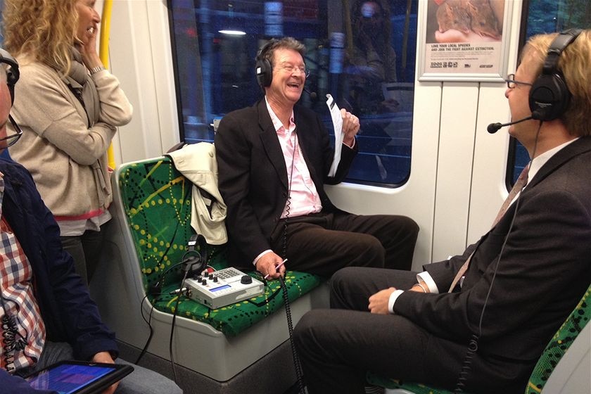 A man laughs as he sits on a tram, wearing a radio headset. Person wearing headset sits opposite, people standing nearby.