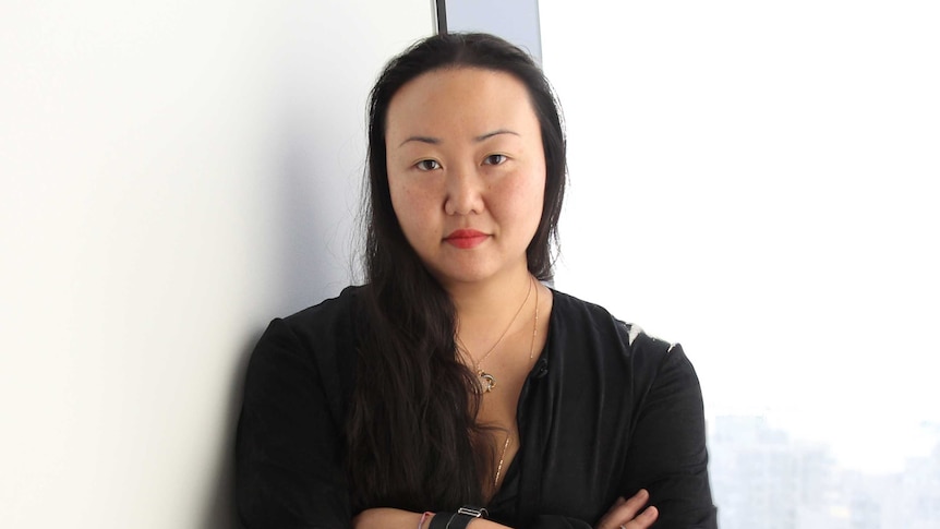Hanya Yanagihara standing with a neutral expression and arms crossed in the corner between a blank cream wall and a window