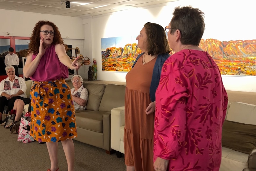 Three women stand acting, while other people sit on couches behind