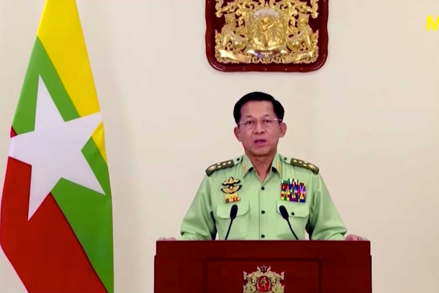 A man in a light green military uniform looks into the camera as he speaks behind a lectern with the Myanmar flag to his right.