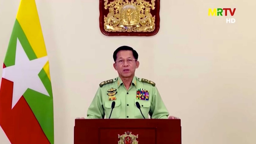 A man in a light green military uniform looks into the camera as he speaks behind a lectern with the Myanmar flag to his right.