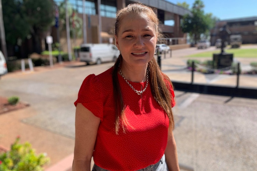 A woman in a red shirt stands outside a building smiling. 