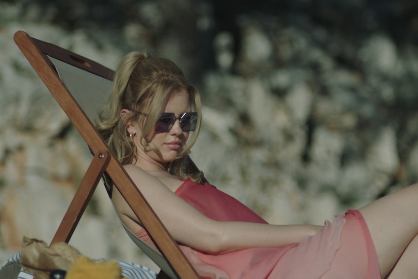 Mia Goth, a white woman with strawberry blonde hair wears a pink caftan and sunglasses, while lounging on the beach