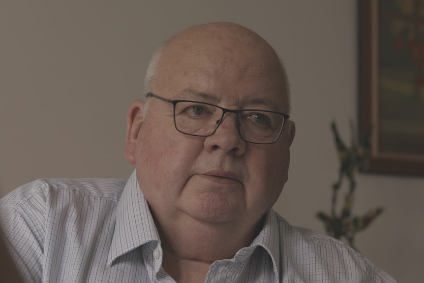 A bald man wearing glasses and a blue striped shirt talking to someone sitting opposite.