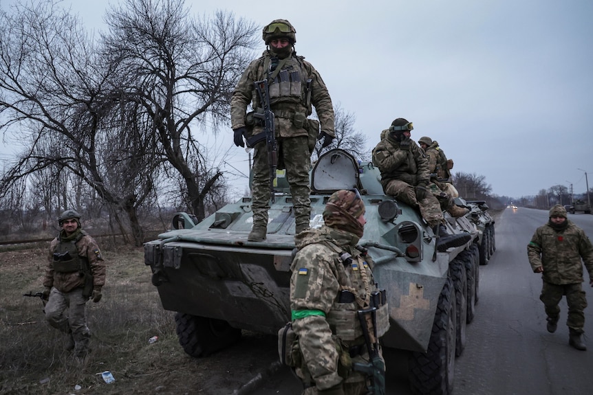 Soldiers stand atop and armoured vehicle that is parked on a long straight road.