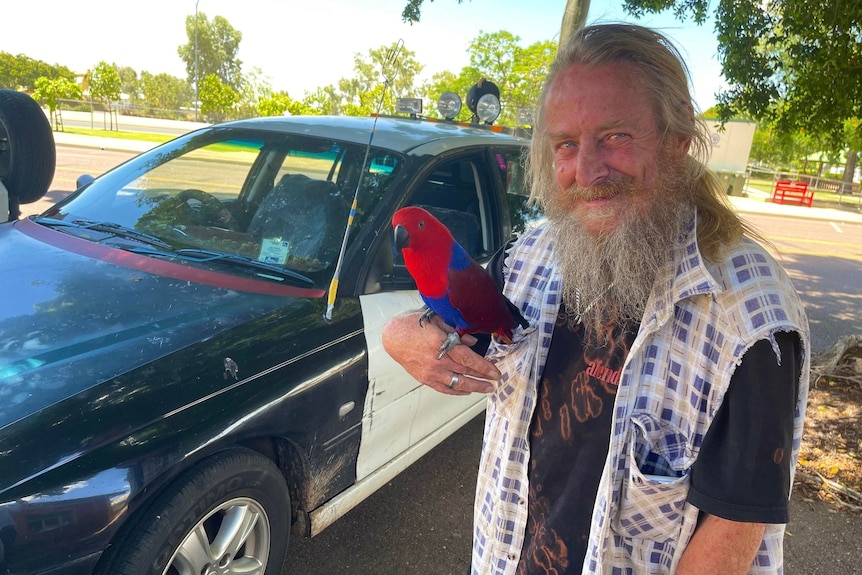 Man standing with red and blue parrot in front of car