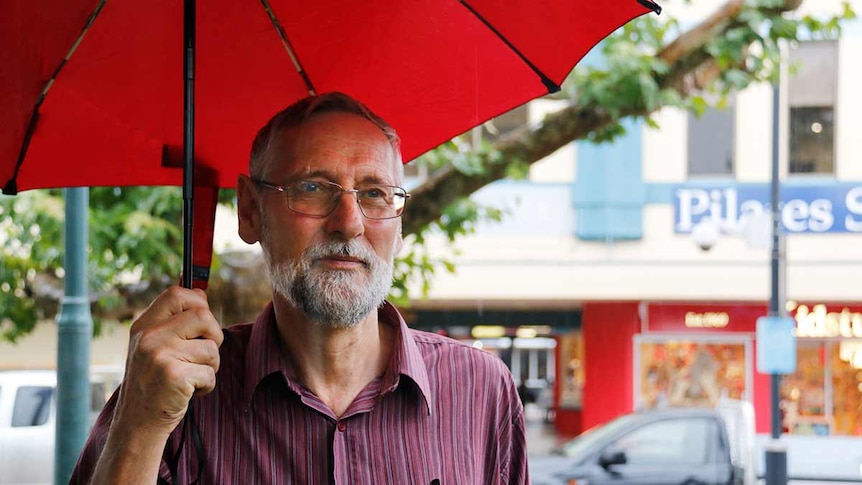 A man stands outside a strip of shops under an umbrella.