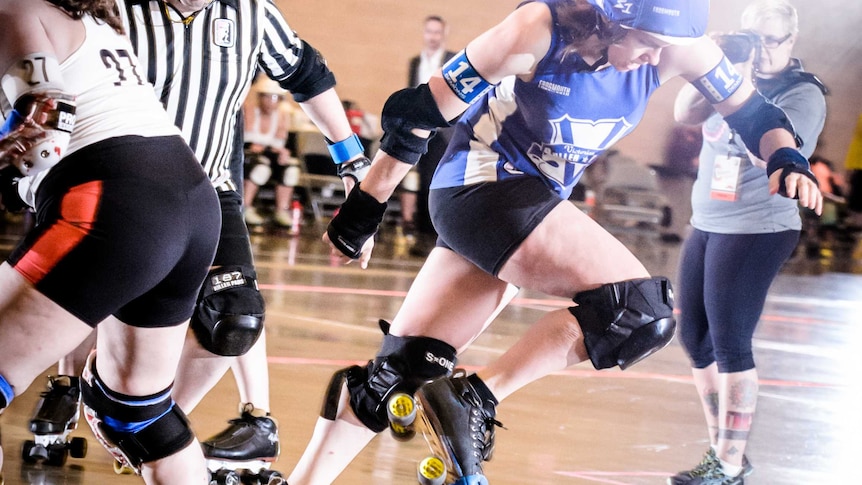 A roller skating woman in blue skips past another in white, referees and photographers in background.
