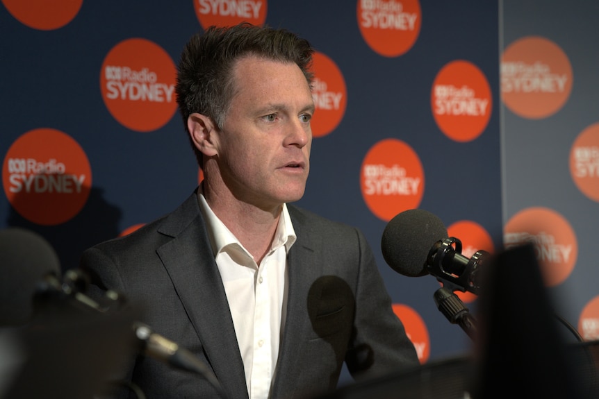 Man in suit in front of microphone with ABC Radio Sydney background behind him