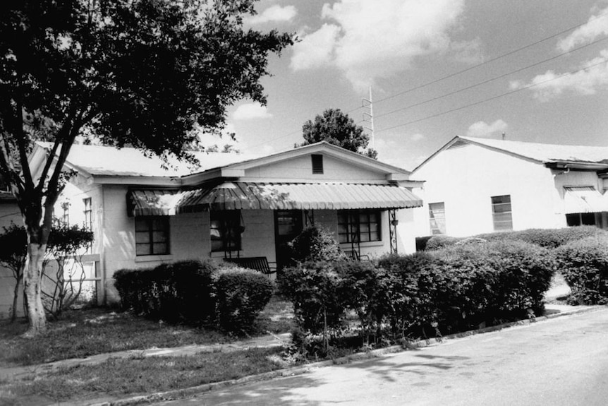 A black and white image of a small house with a hedge and tree in the front yard