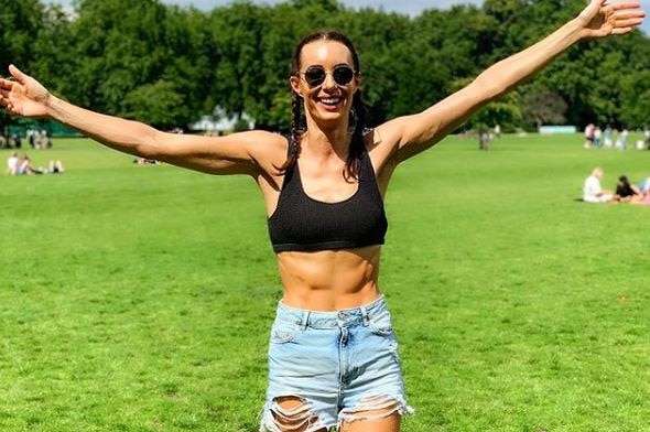 Emily Hartridge smiles as she walks forwards with her arms open wide. She wears a black sports bra and denim shorts.