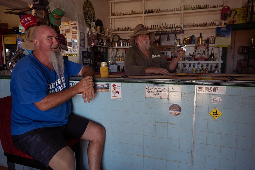 A man sits on a stool in a quirky outback pub, the publican wears a big hat behind the bar
