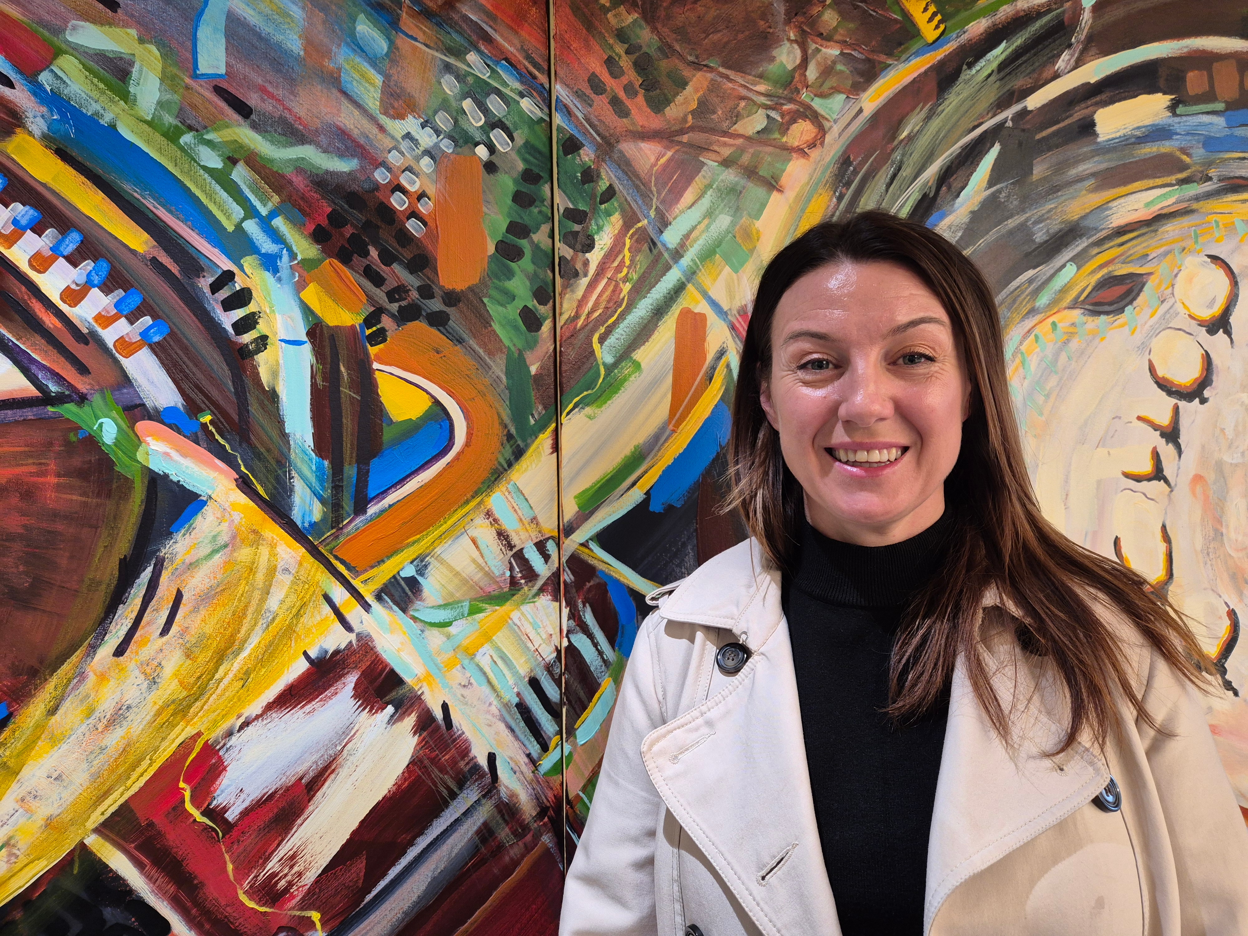A brown haired woman stands in front of a multi-coloured mural, smiling