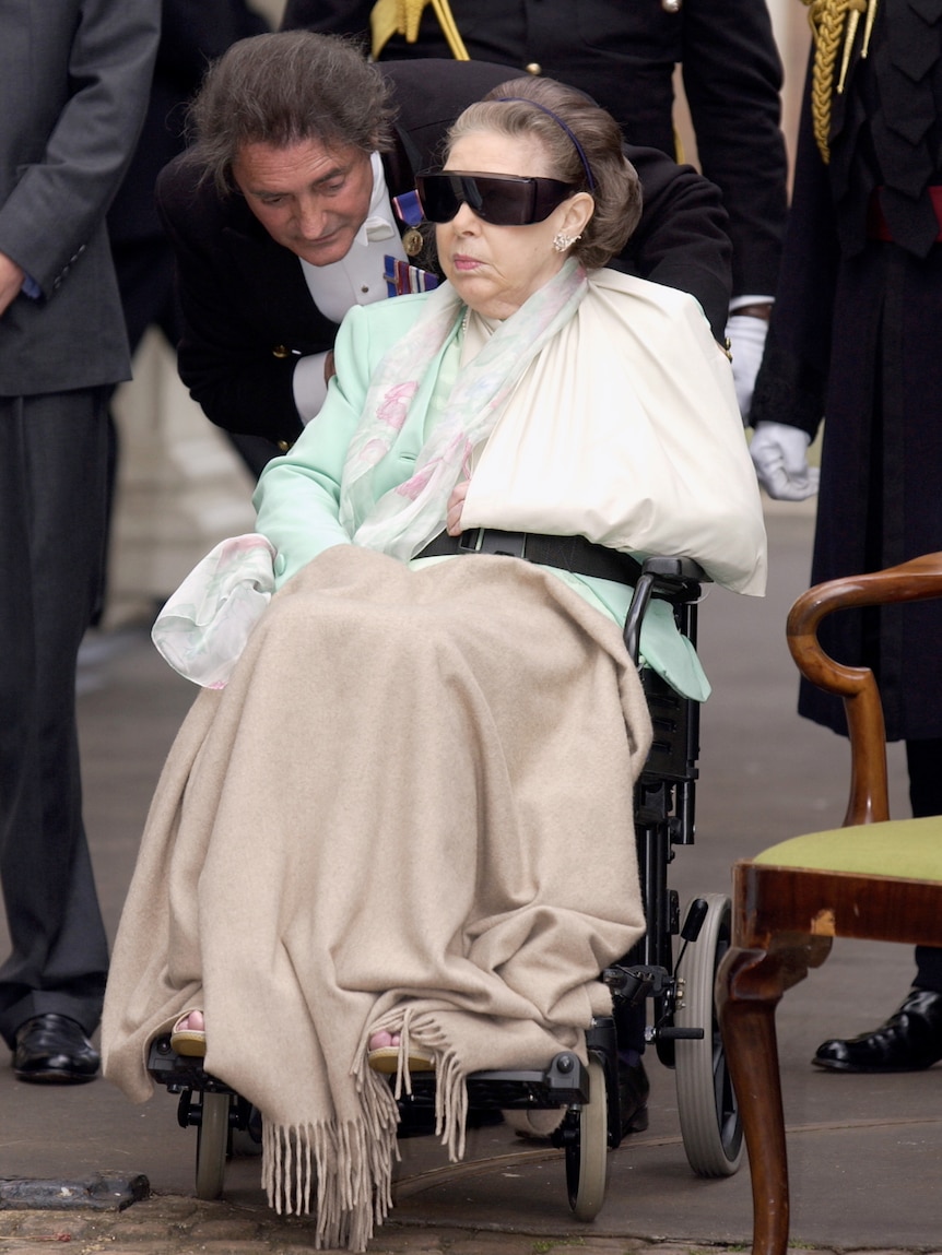Princess Margaret sits in a wheelchair, wearing dark sunglasses, floral scarf and a blanket over her legs. One arm is in a sling