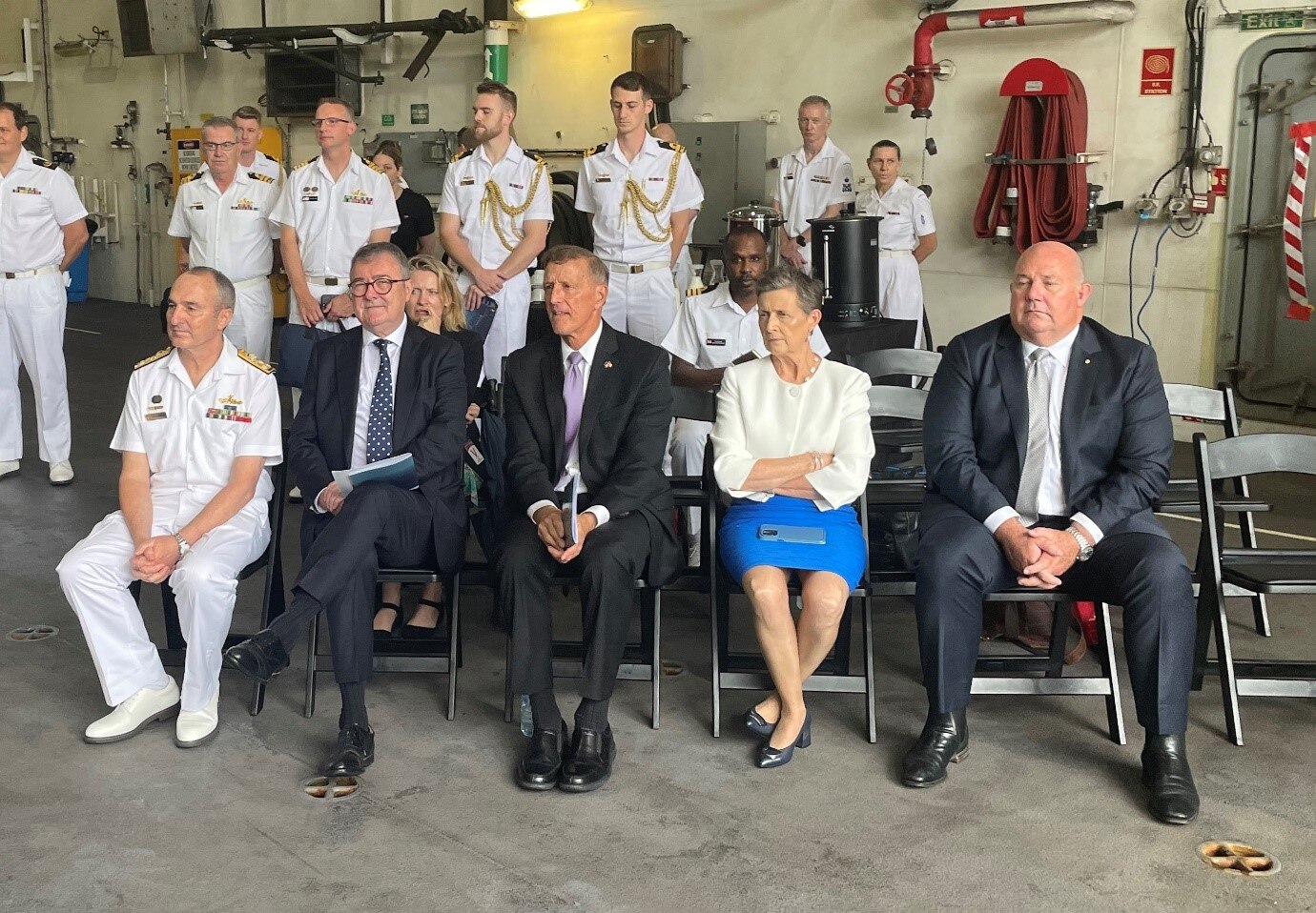 Five people in suits and navy attire sit in a row on chairs, more navy members behind them.
