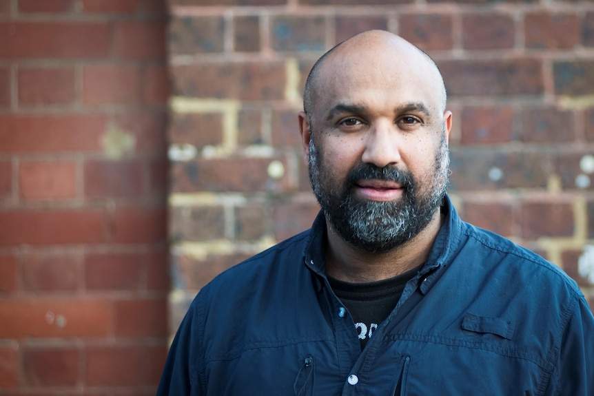 A black man wearing a blue shirt with beard, standing in front of a brick wall