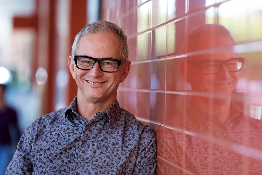 A smiling man in thick-rimmed spectacles leaning up against a tiled wall.