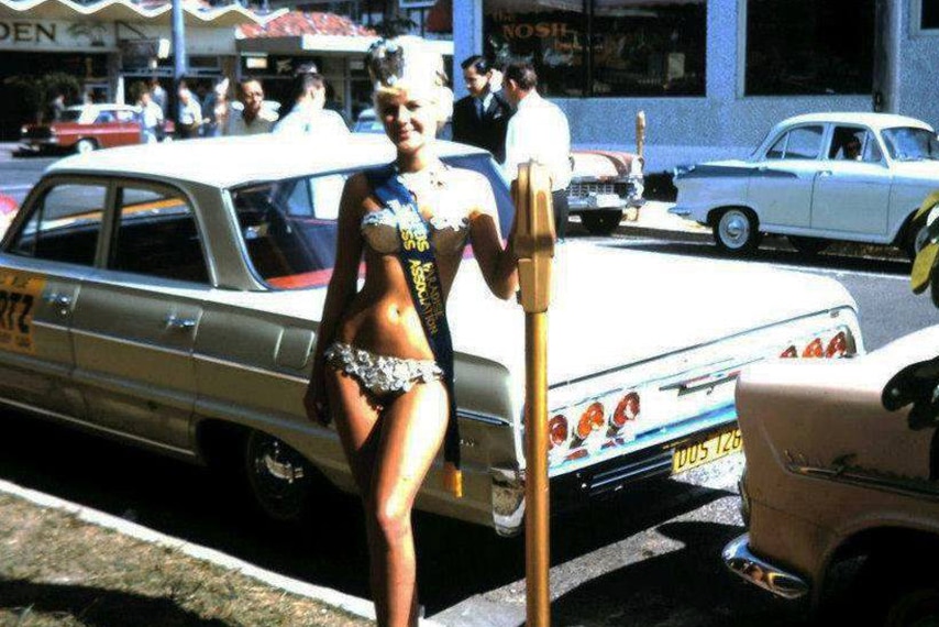 A woman dressed in a gold lame bikini in the 1960s standing in front of a parking meter in street