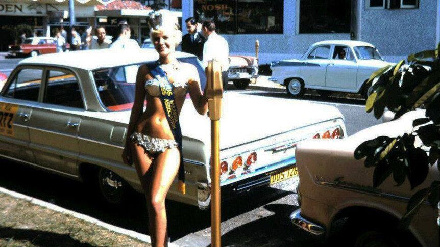 A woman dressed in a gold lame bikini in the 1960s standing in front of a parking meter in street