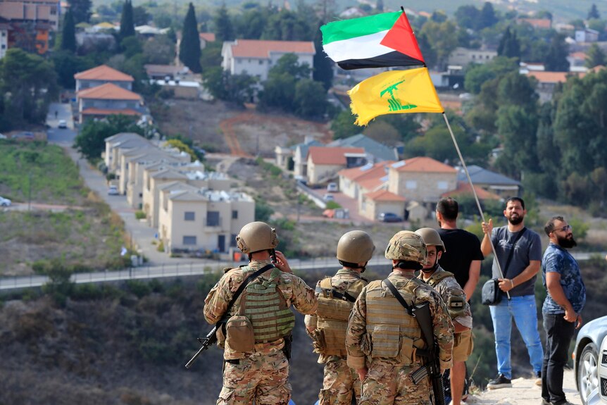 Four soldiersstand on top of a hill looking down on a village, a man in front of them holds the Hezbollah and Palestinian flags