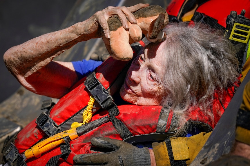 An elderly woman clutches a teddy as she wears a life vest and is rescued by firefighters