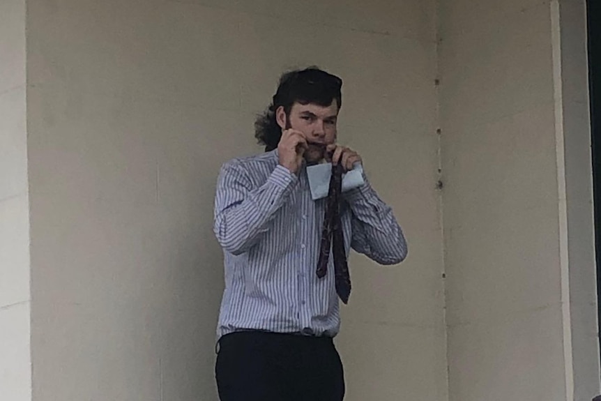 A man stands against a wall with something in his mouth, wearing a blue button-up shirt.