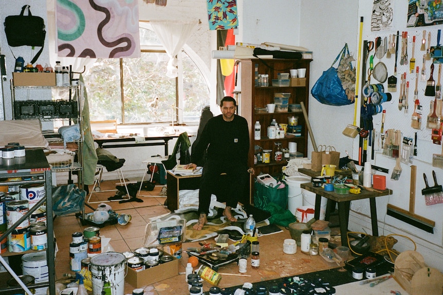A man dress in black sits on a bench, he is surrounded by artworks and art supplies. 