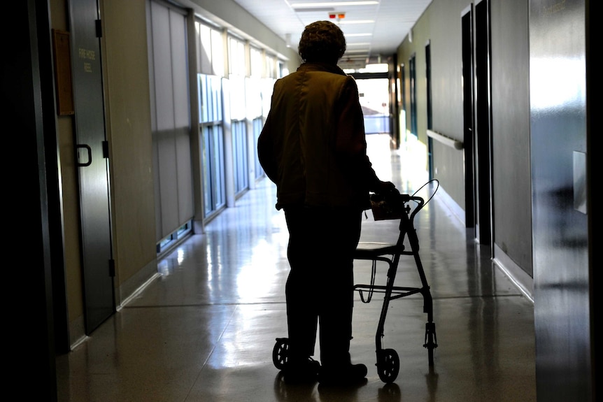 A man in shadow leans on a walker as he faces away from camera in a corridor inside a group home
