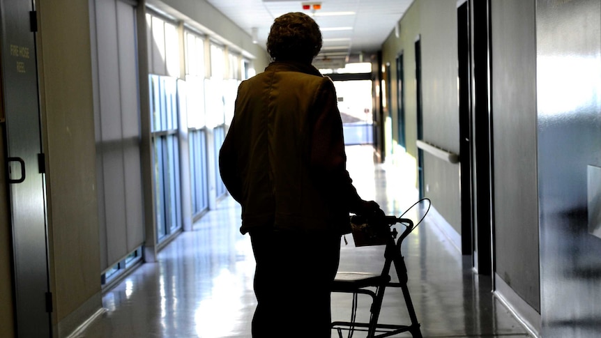 A man in shadow leans on a walker as he faces away from camera in a corridor inside a group home