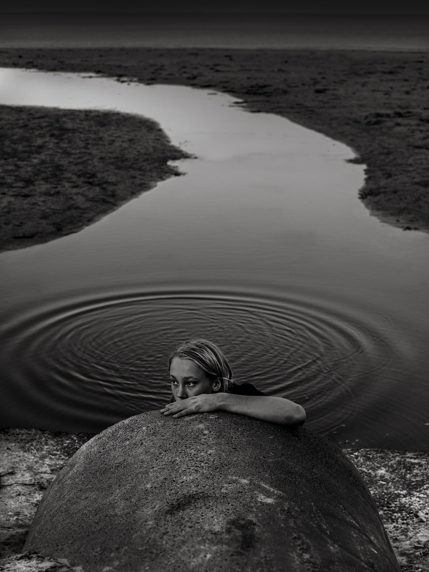 The photo is black and white and of a child leaning on a drain near water.