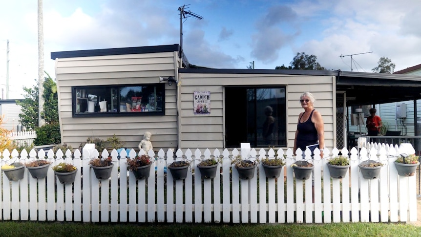 A woman standing in front of a small house with a white picket fence with hanging flower baskets