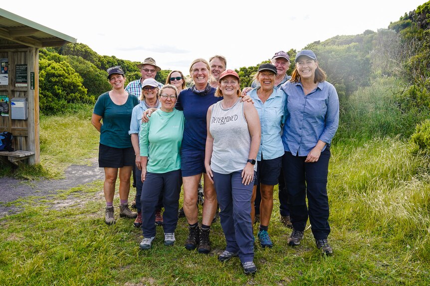 A group of walkers stand in a grassy campsite with their arms around each other, smiling.
