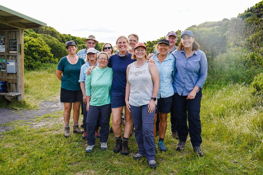 A group of walkers stand in a grassy campsite with their arms around each other, smiling.