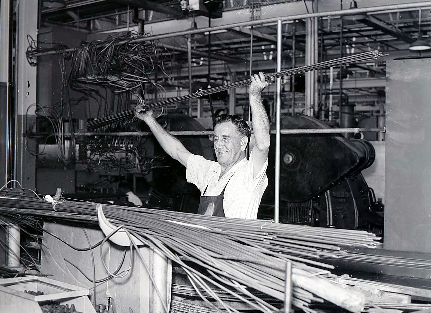 A black and white photograph of a man holding metal tubing above his head in a factory