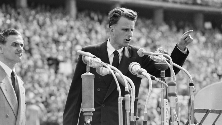 Black and white image of a young Billy Graham pointing while addressing a large crowd.