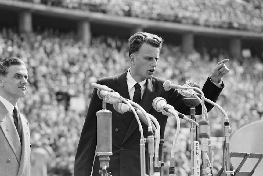 Black and white image of a young Billy Graham pointing while addressing a large crowd.