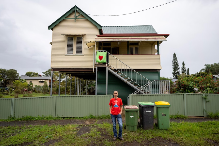 Jo standing on damp ground in front of her home, a wooden house on stilts with a mural of a heart hanging from the stairs.