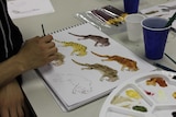 A hand holding a paintbrush rests on a book covered with half-painted lion-like animals.