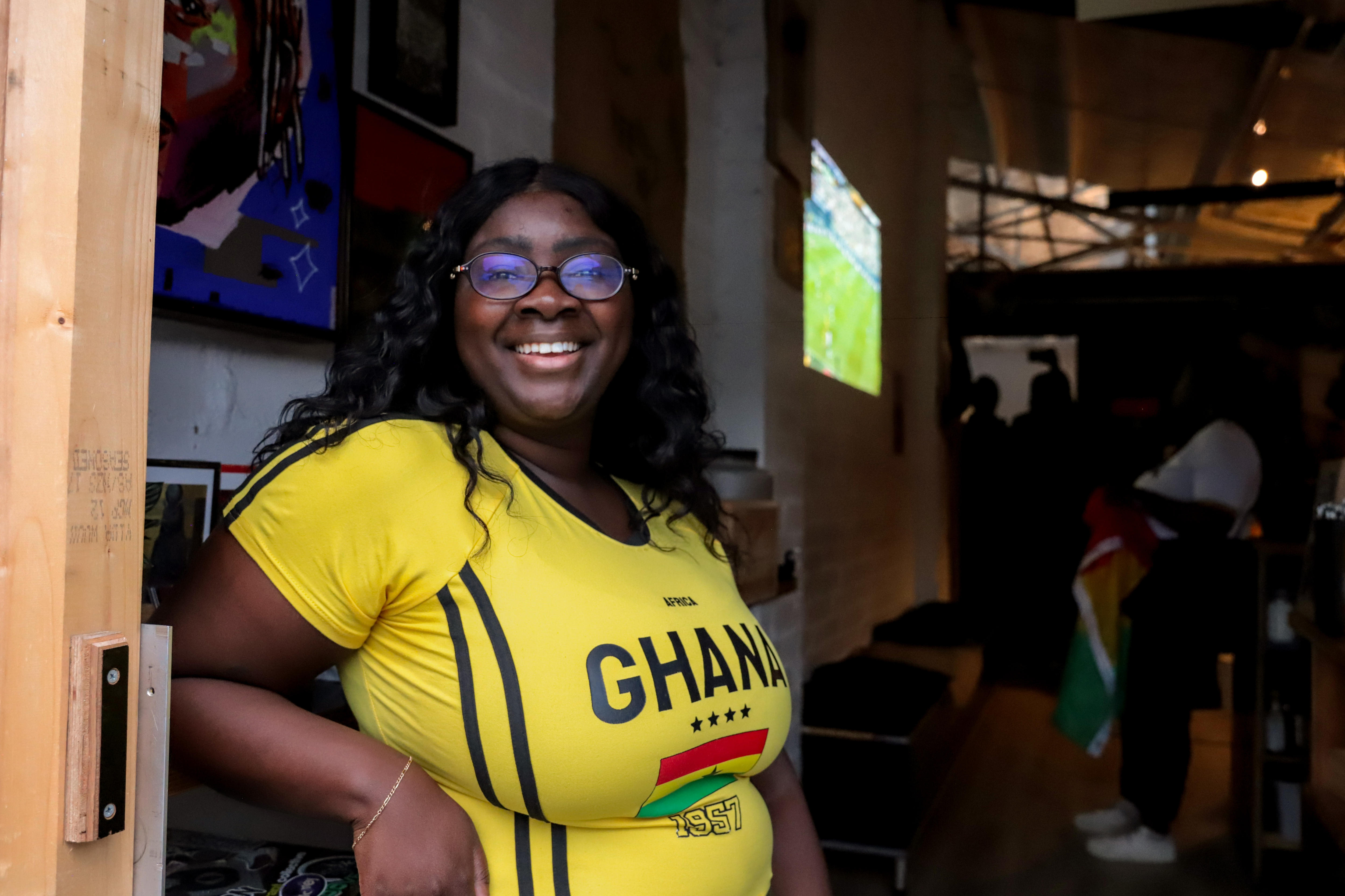 A woman wearing a yellow t-shirt with GHANA written on the front stands with a large tv screen and crowd of people behind.