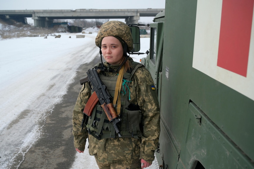 Une femme en tenue de camouflage militaire se tient à côté d'un véhicule médical. 