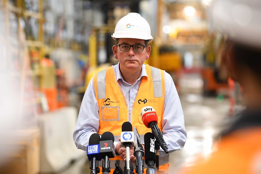 Daniel Andrews wears a hard hat and a high-vis vest as he stands in front of media microphones.