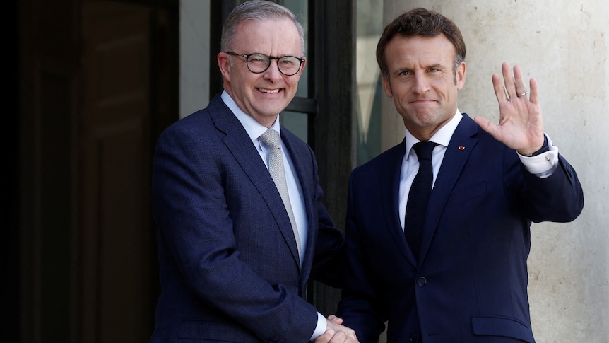 Two men shake hands for camera as one waves.