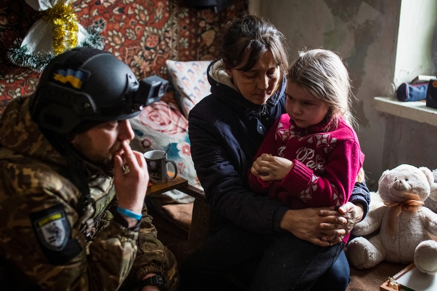 A policeman kneels to comfort a little girl sitting on her mother's lap surrounded by cuddly toys.