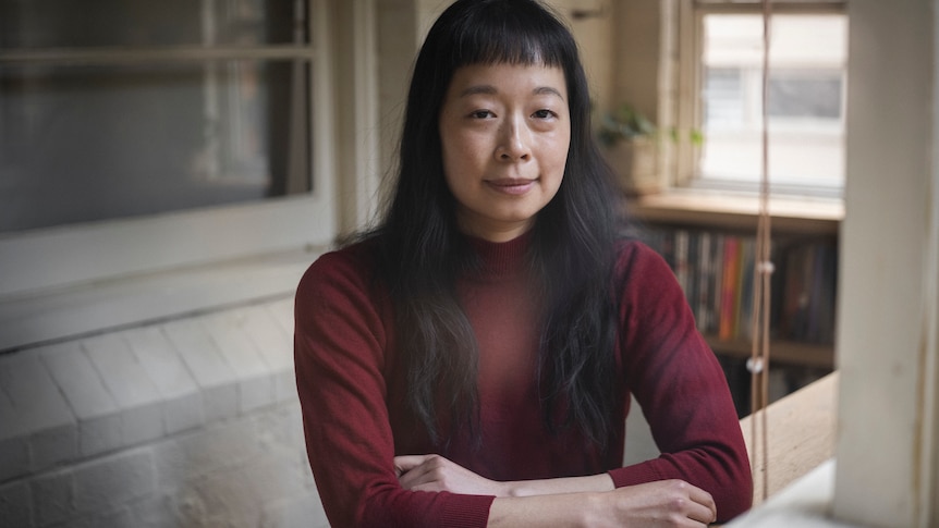 A woman with black hair sits with her arms folded at a table inside a home.