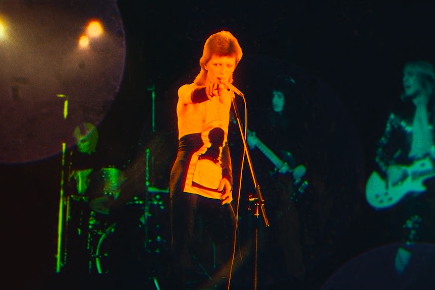David Bowie shirtless and with a mullet sings into a microphone lit in orange against a blue and green lit stage.