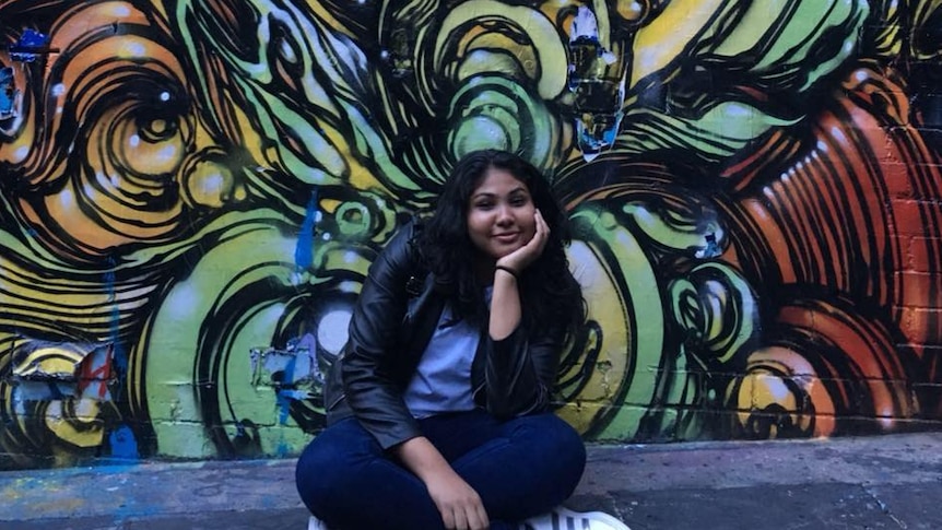 Nisali Perera sits in front of a graffiti art wall smiling.