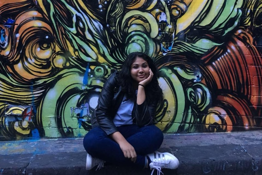 Nisali Perera sits in front of a graffiti art wall smiling.