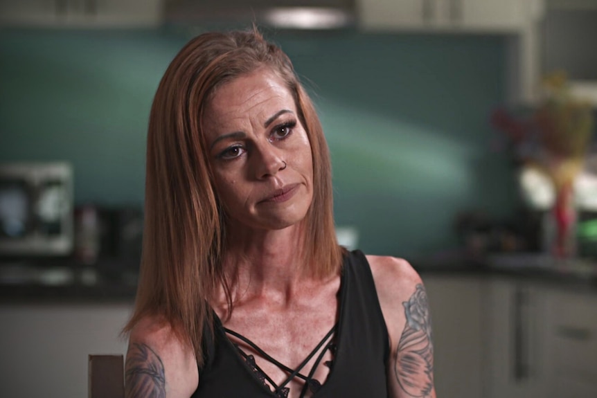 Woman with shoulder-length red hair and tattoos on her arms, sitting in a kitchen. 